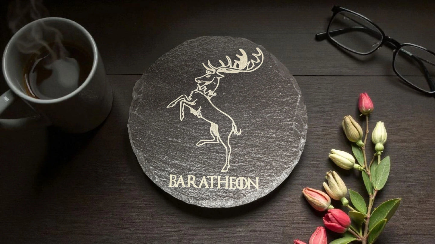Slate coaster with a deer design and 'Baratheon' text on a wooden surface with a mug, glasses, and flowers.