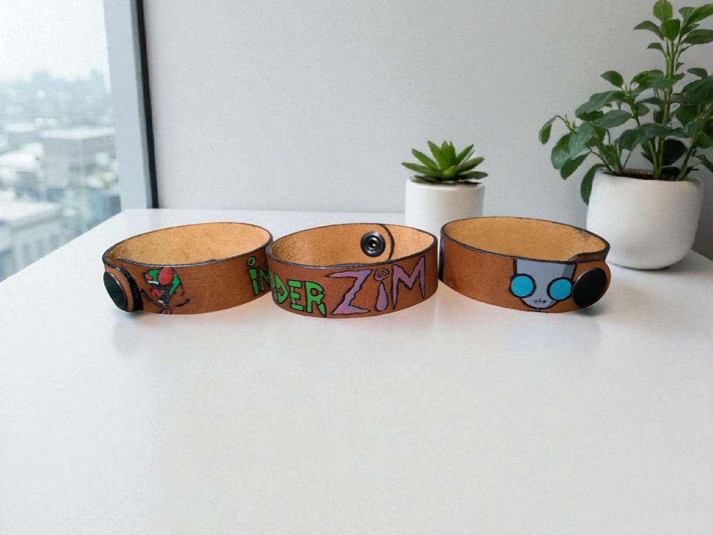 Three InvaderZim leather bracelets with unique designs on a white surface with a cityscape view.