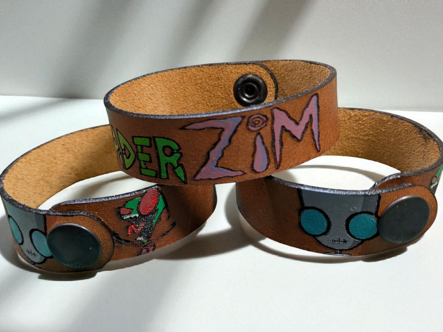 Invader Zim themed full grain leather bracelets sat on a off white worktop with light shinning down on them.