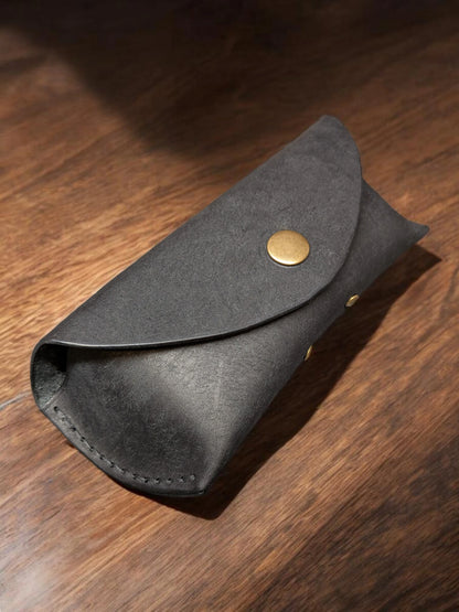 Black Badalassi Pueblo leather sunglasses case with an envelope flap and brass snap, on a wooden table