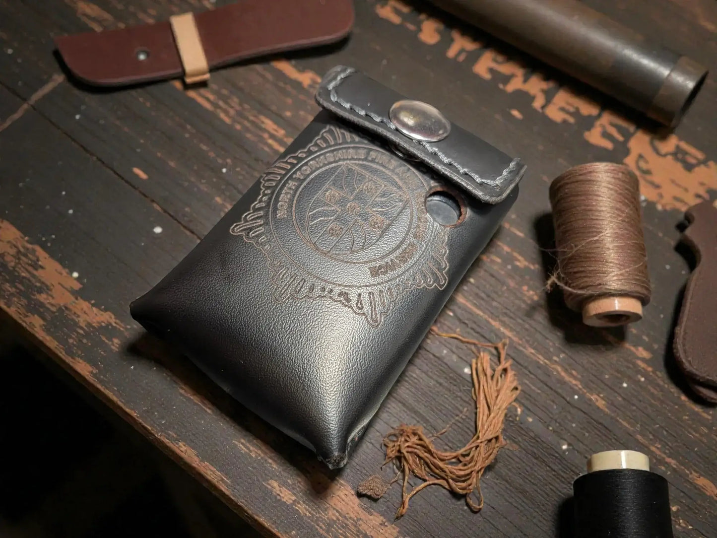 Black leather Alerter case with embossed North Yorkshire fire and rescue design on a wooden surface with thread and other materials.
