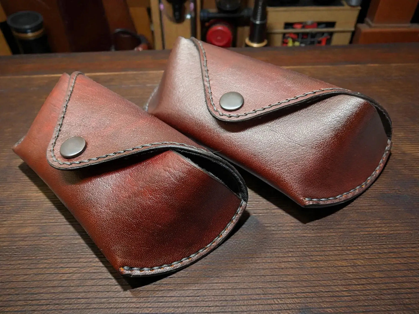Two brown full grain leather sunglasses cases on a wooden surface with a workshop background.