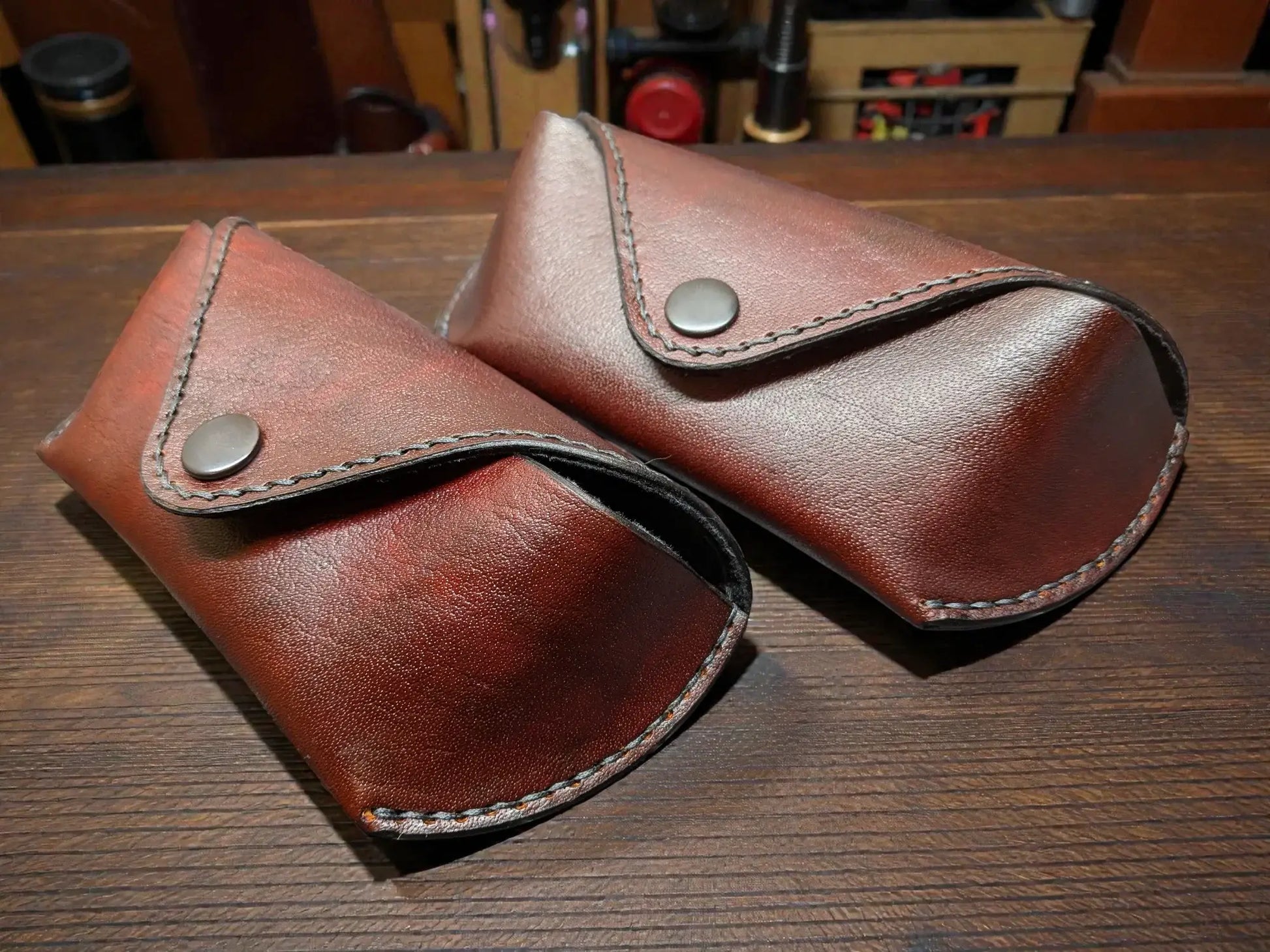 Two brown full grain leather sunglasses cases on a wooden surface with a workshop background.