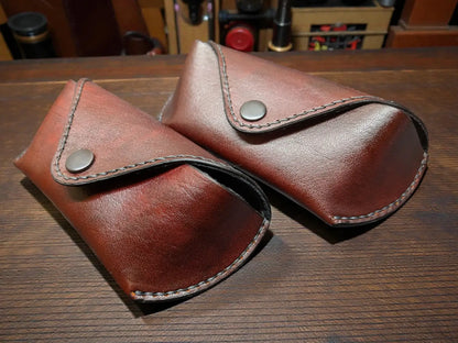 Two brown full grain leather sunglasses cases on a wooden surface with a workshop background.