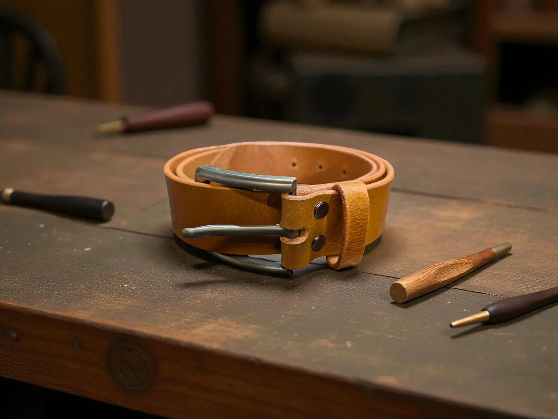 Tan leather belt with metal buckle on a wooden surface with tools.