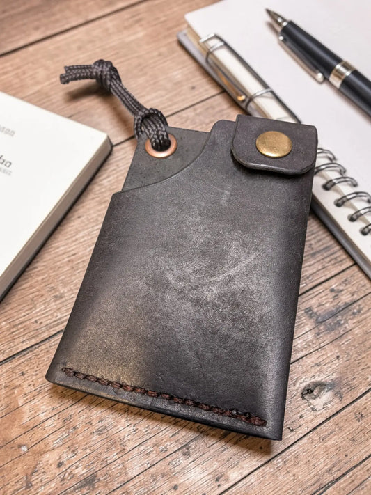 Minerva' Compact Card Holder in Badalassi Pueblo Leather - Handmade Minimalist Wallet shown in dark vegetable tanned leather with brass button closure, resting on wooden desk with notebooks and pen nearby.