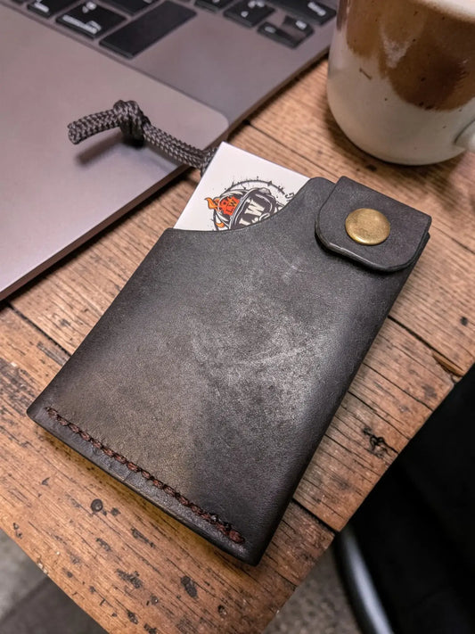 Minerva' Compact Card Holder in Badalassi Pueblo Leather - Handmade Minimalist Wallet resting on a wooden desk next to a laptop and coffee mug, showcasing its rugged full grain leather texture and brass snap closure.