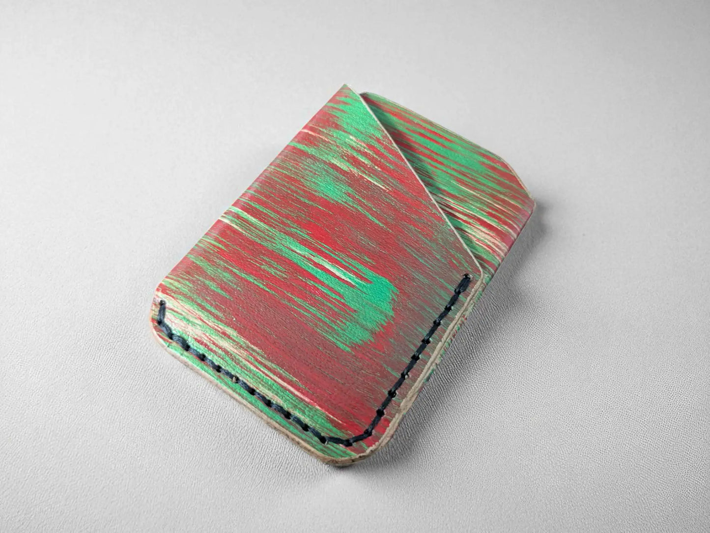 Red and green splash-dyed vegetable tanned leather card wallet, closed, front view on a grey surface