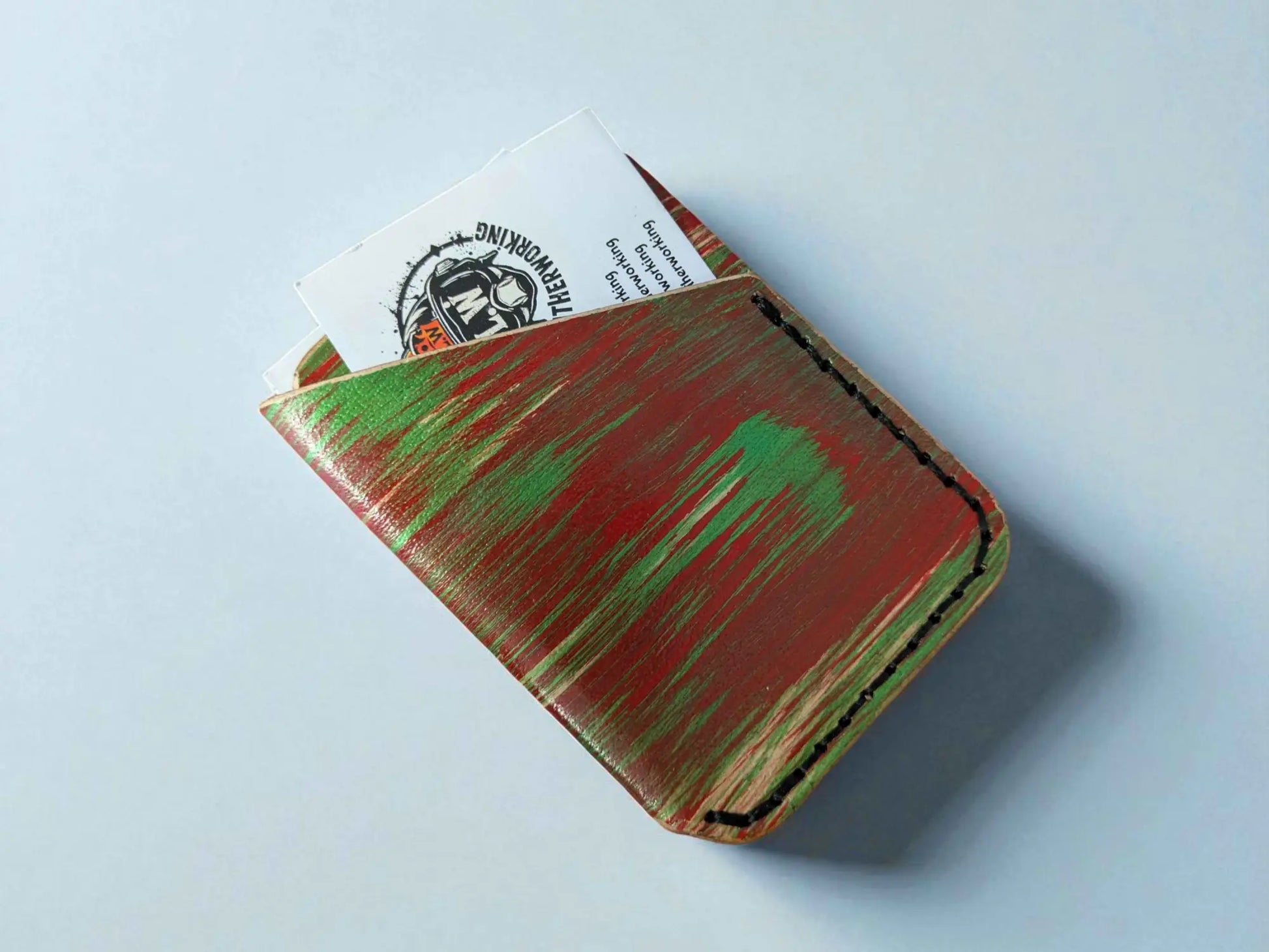 Red and green splash-dyed vegetable tanned leather card wallet with two cards peeking out, on a light grey surface