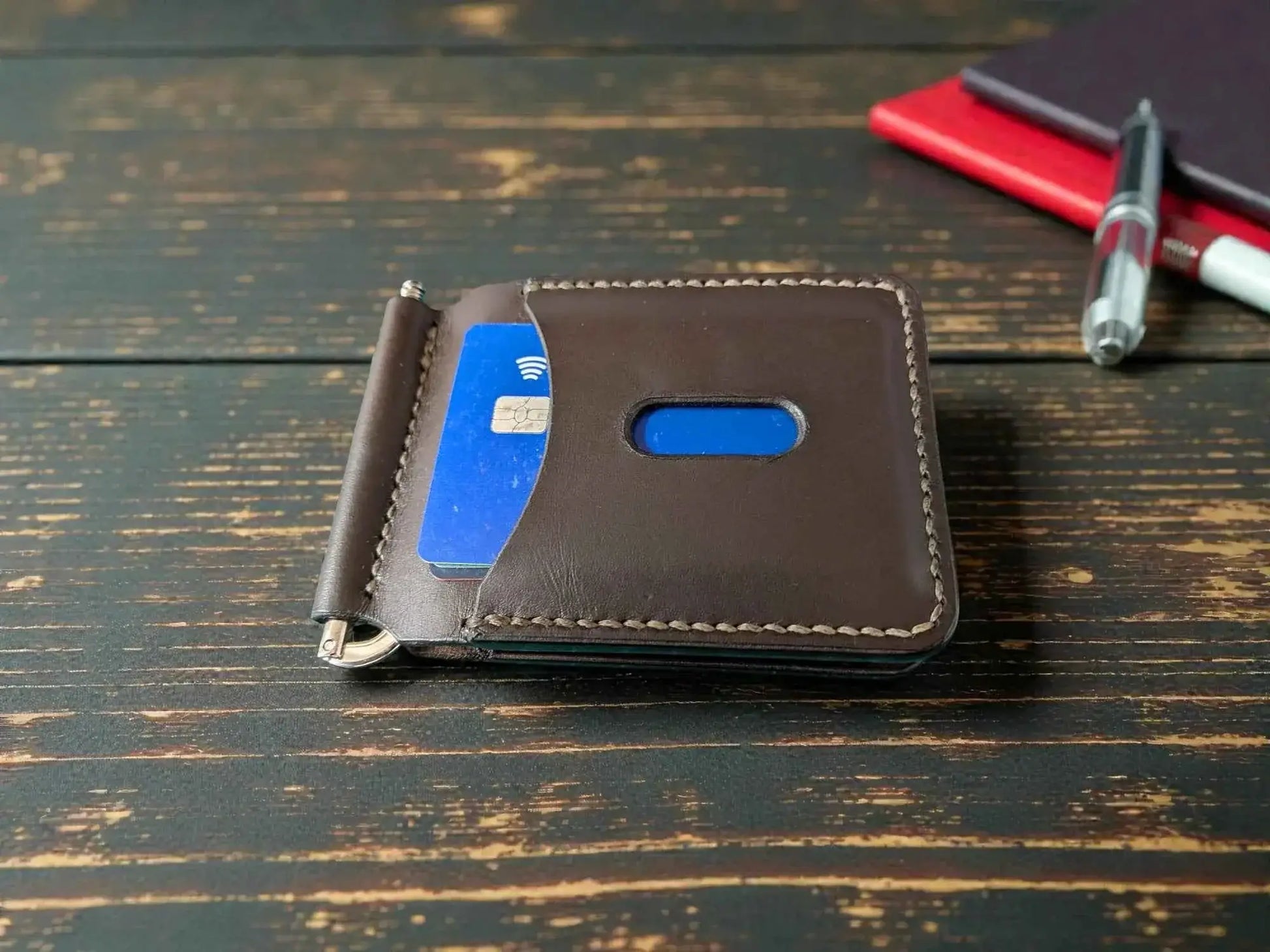 Patriot' Money Clip Wallet - Handmade Minimalist Leather Wallet in dark brown full grain leather with card partially inserted, on rustic wooden surface with pen and notebook in background.