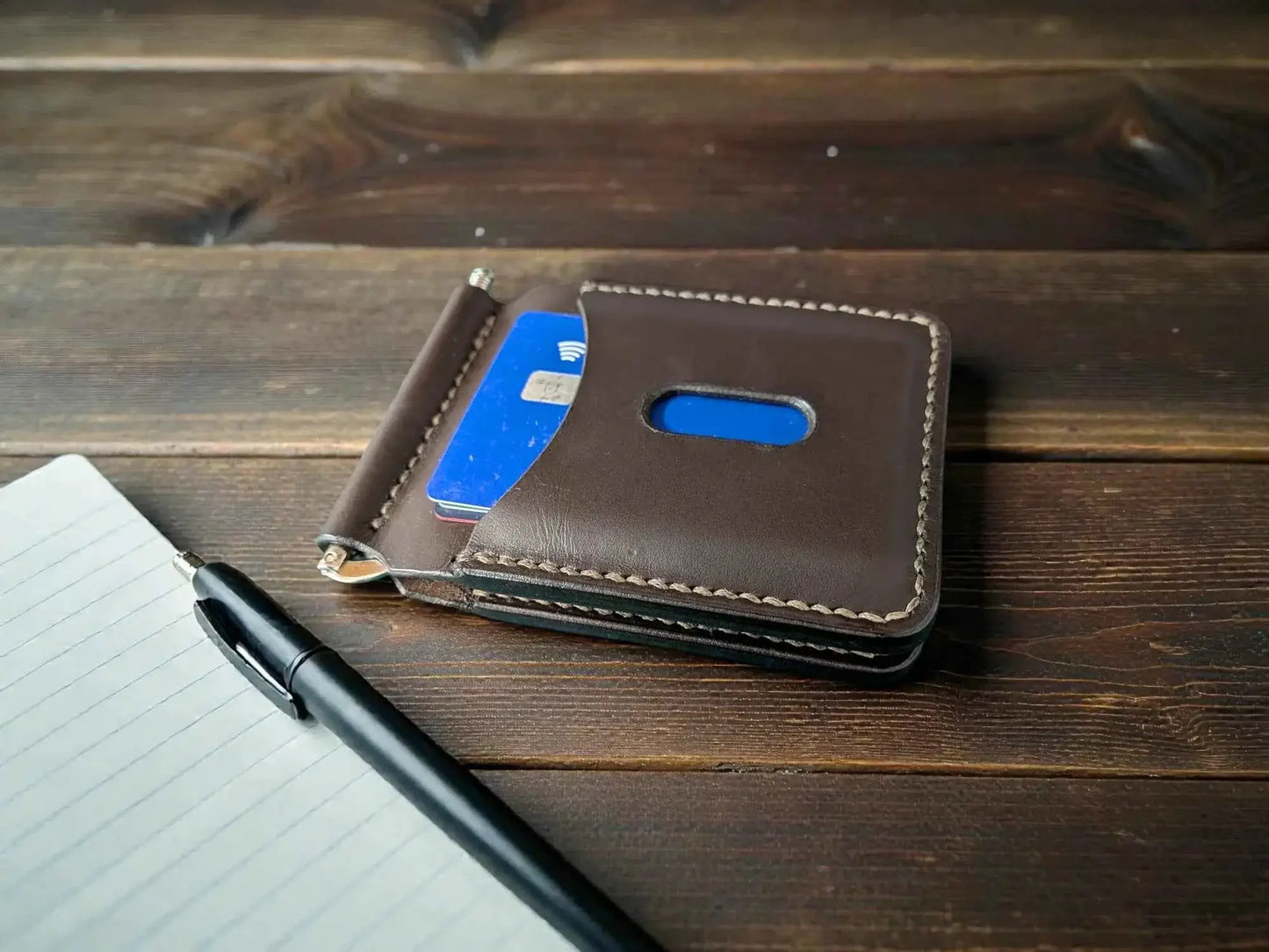 Patriot' Money Clip Wallet - Handmade Minimalist Leather Wallet in dark brown full grain leather, holding blue cards, positioned on a wooden surface next to a pen and notebook.