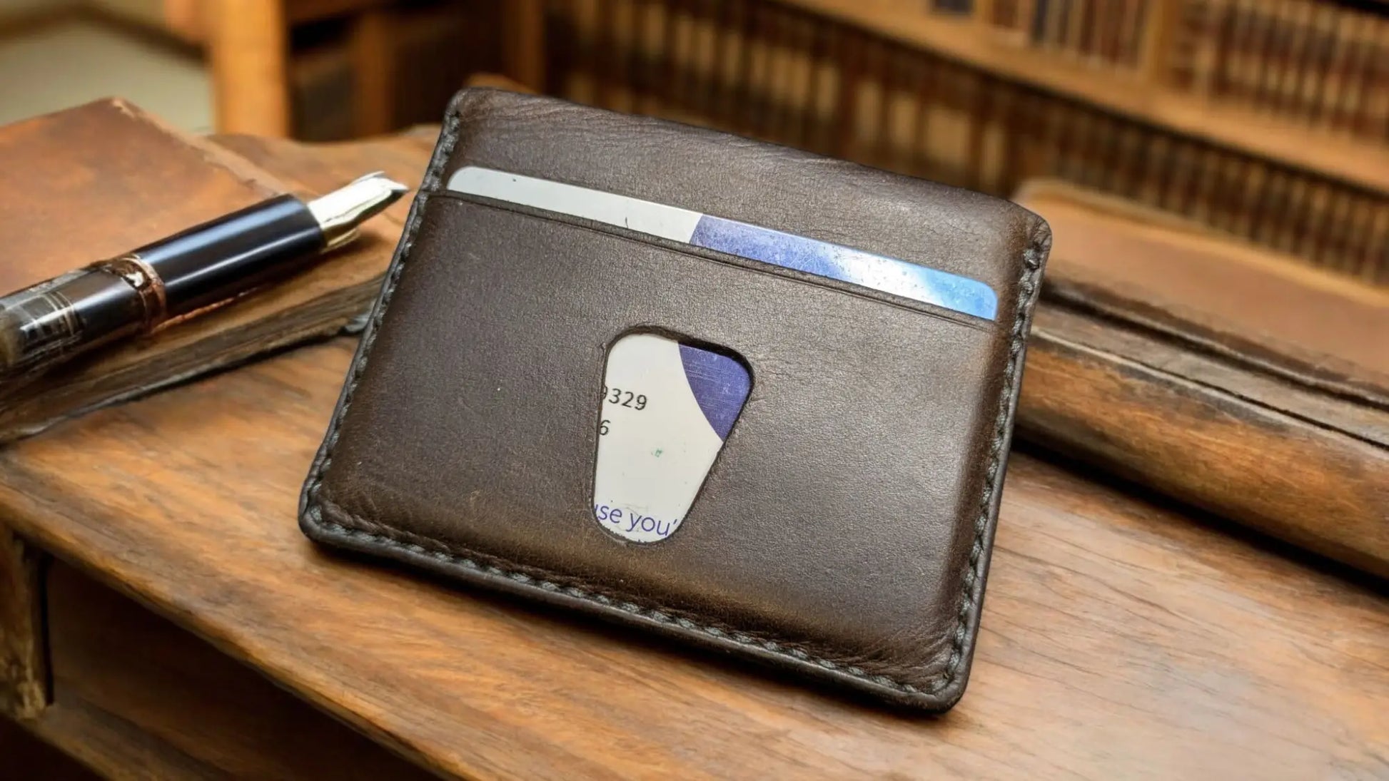 Brown leather Solaris minimalist card holder holding a bank card, on a wooden desk with library shelves in the background